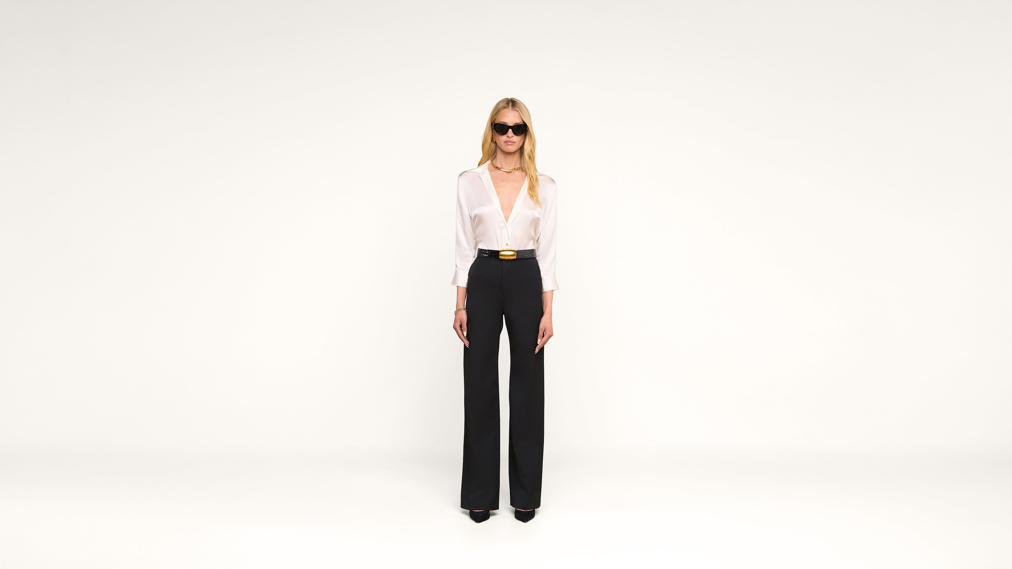 A woman wearing sunglasses, a white blouse, and high-waisted black pants stands against a plain white background.