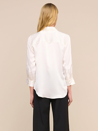 Una persona con el pelo largo y rubio, vista de espaldas, lleva la blusa Dani Silk en color marfil, combinada con unos pantalones negros, sobre un fondo liso de color beige.