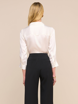 Una mujer con el pelo largo y rubio, vista de espaldas, lleva la blusa Dani Silk en color marfil —con las mangas remangadas de forma desenfadada— combinada con unos pantalones negros de cintura alta, sobre un fondo beige.