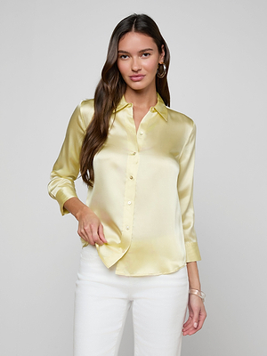 A woman with long brown hair stands against a plain light gray background, wearing the shiny yellow Dani Silk Blouse and white pants, looking at the camera with a slight smile.