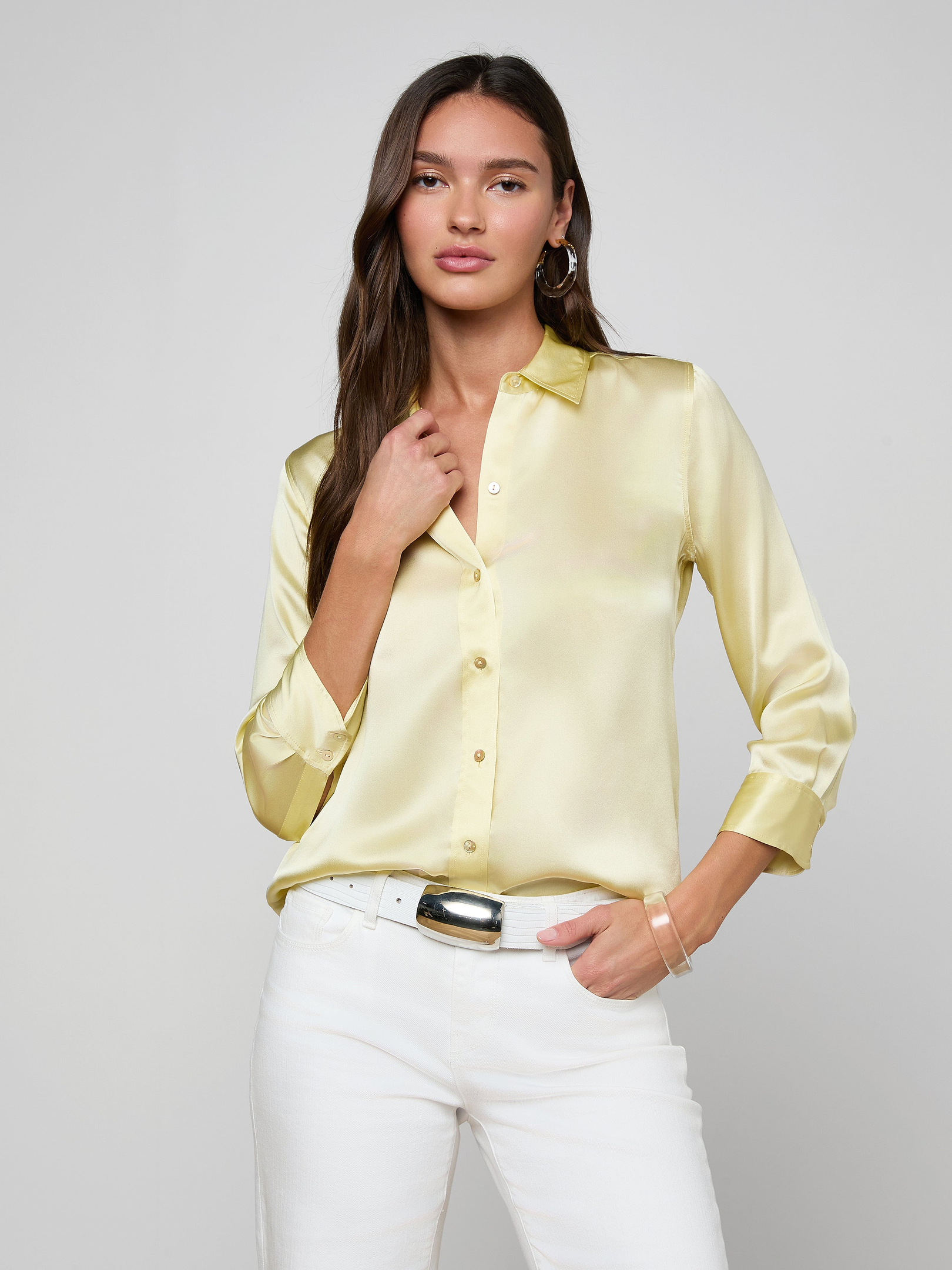 A woman wearing the Dani Silk Blouse in light yellow and white pants stands against a plain light background, with one hand touching her collar and the other in her pocket.