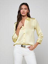 A woman wearing the Dani Silk Blouse in light yellow and white pants stands against a plain light background, with one hand touching her collar and the other in her pocket.