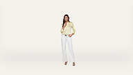 Woman standing against a plain light background, wearing a shiny light yellow blouse, white pants, and heeled sandals. She has one hand in her pocket and is smiling gently at the camera.