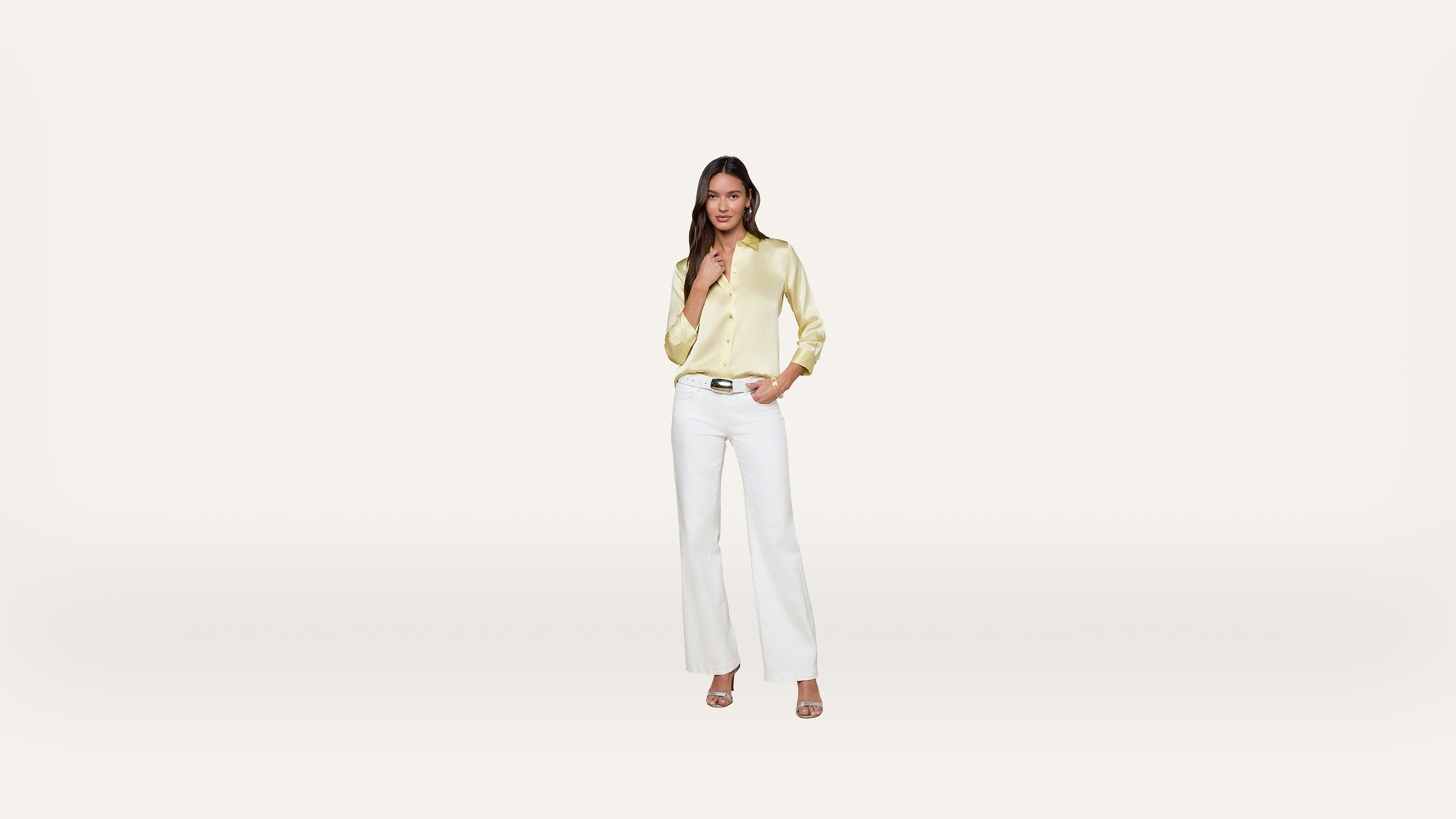 Woman standing against a plain light background, wearing a shiny light yellow blouse, white pants, and heeled sandals. She has one hand in her pocket and is smiling gently at the camera.
