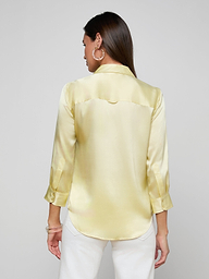 A woman with long brown hair is shown from the back against a plain light background, wearing the Dani Silk Blouse—a light yellow satin top with three-quarter sleeves—and white pants.