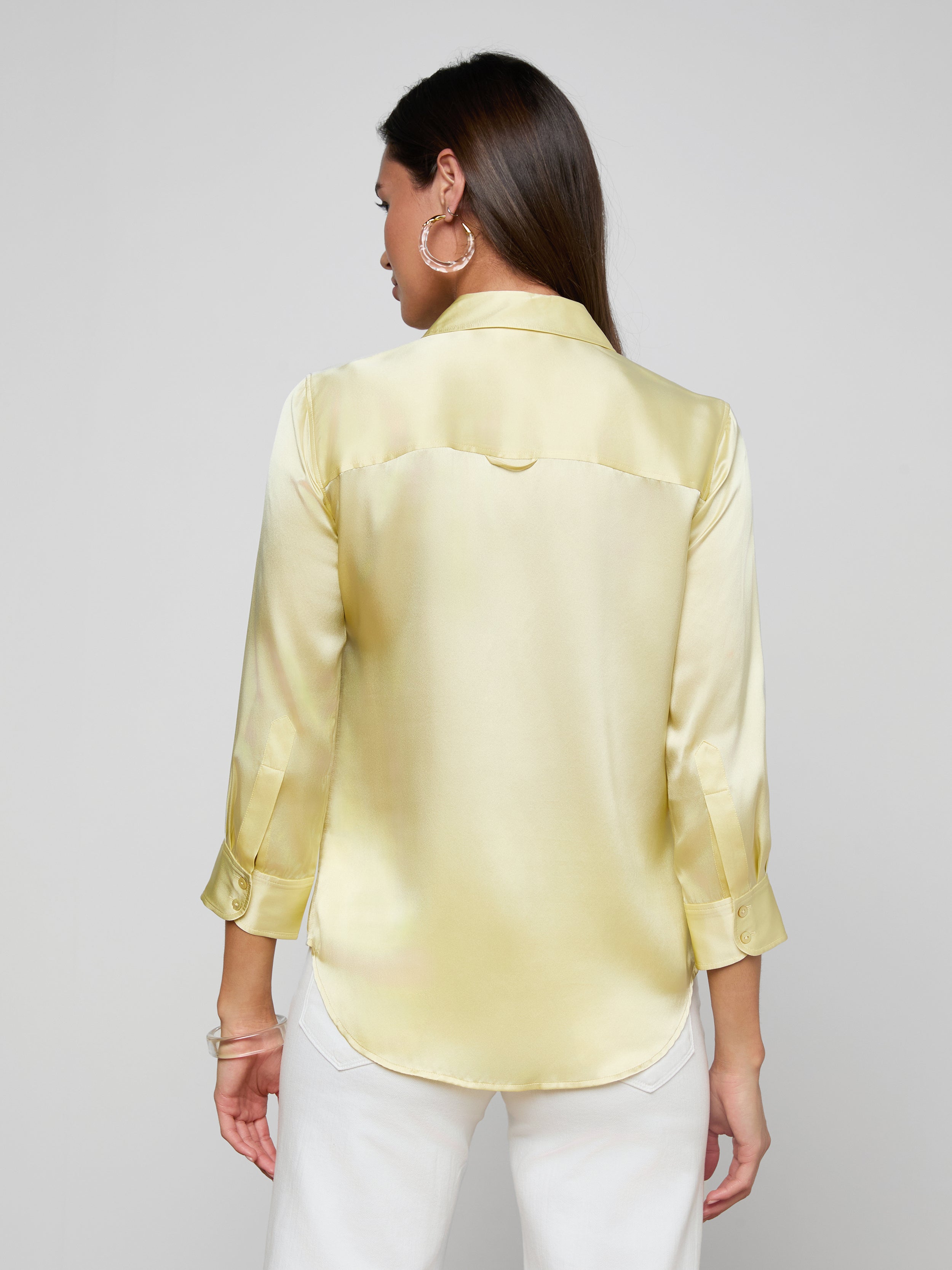 A woman with long brown hair is shown from the back against a plain light background, wearing the Dani Silk Blouse—a light yellow satin top with three-quarter sleeves—and white pants.