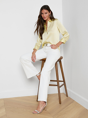 A woman with long brown hair sits on a wooden stool in a bright indoor setting, wearing the Dani Silk Blouse in light yellow, paired with white pants and heeled sandals. Light walls and wood flooring complete the scene.