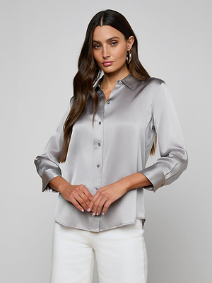 A woman with long brown hair wears the Dani Silk Blouse—a shiny silver, long-sleeve button-down—paired with white pants and hoop earrings, standing against a plain light gray background with a neutral expression.
