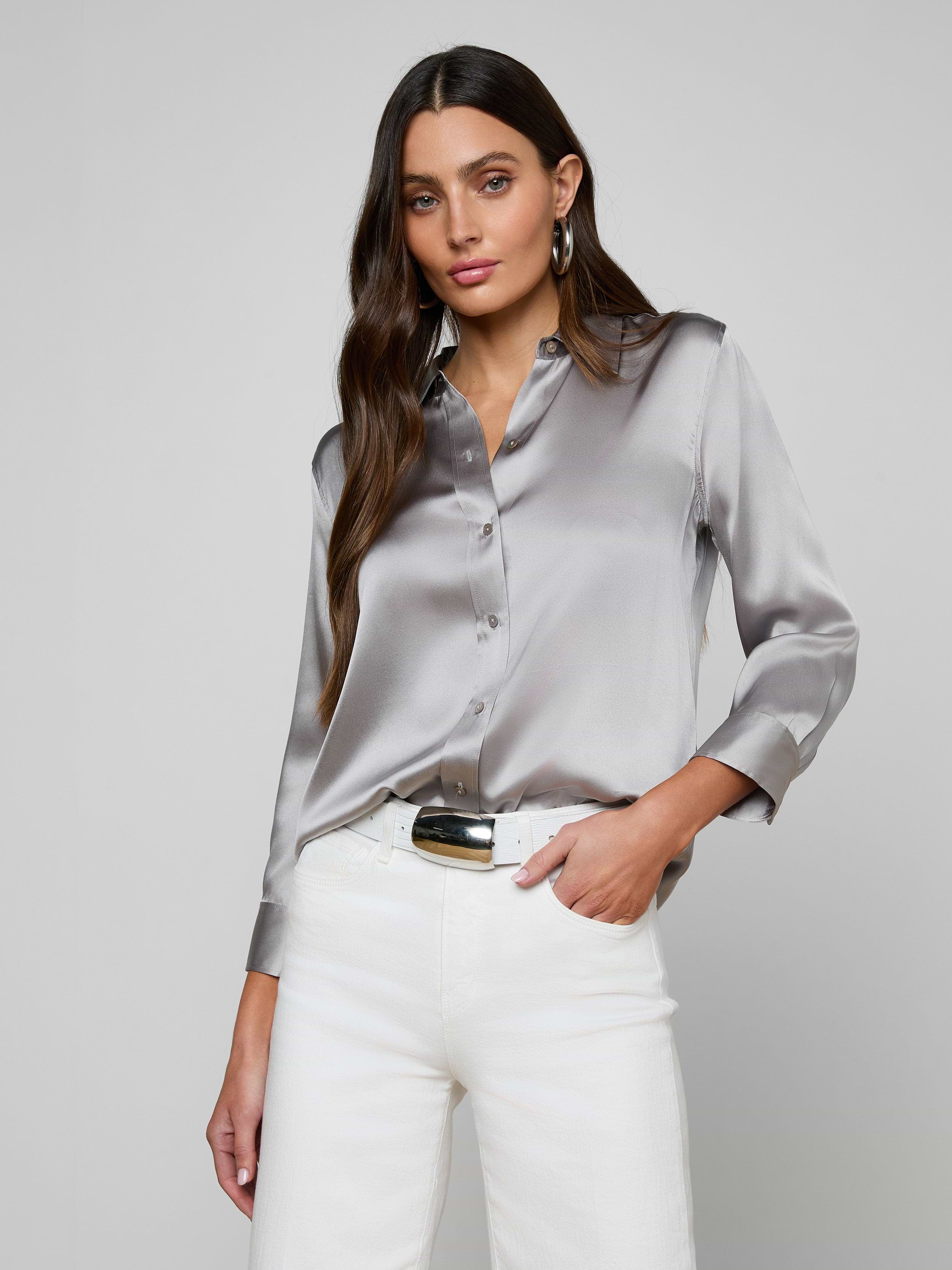 A woman wearing the Dani Silk Blouse, a shiny long-sleeved button-down top, paired with white pants, stands with one hand in her pocket against a plain, light background.