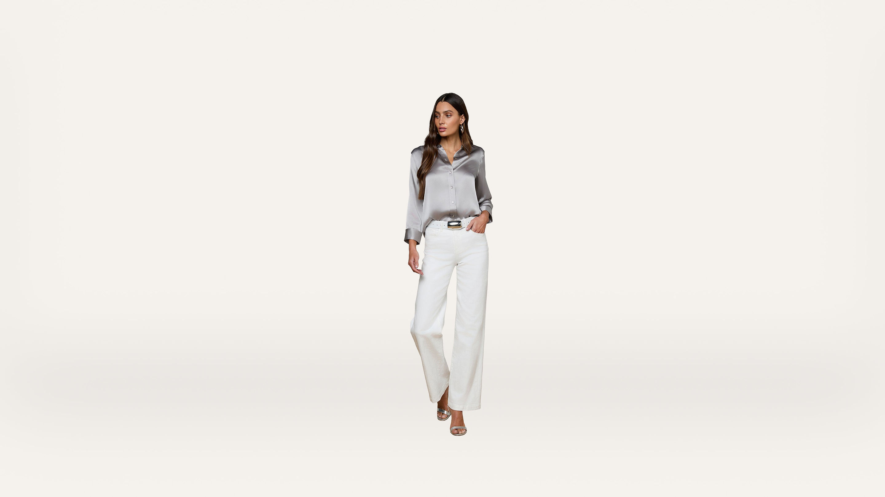 A woman wearing a shiny silver blouse, white wide-leg pants, and beige heels stands against a plain white background, looking to the side.