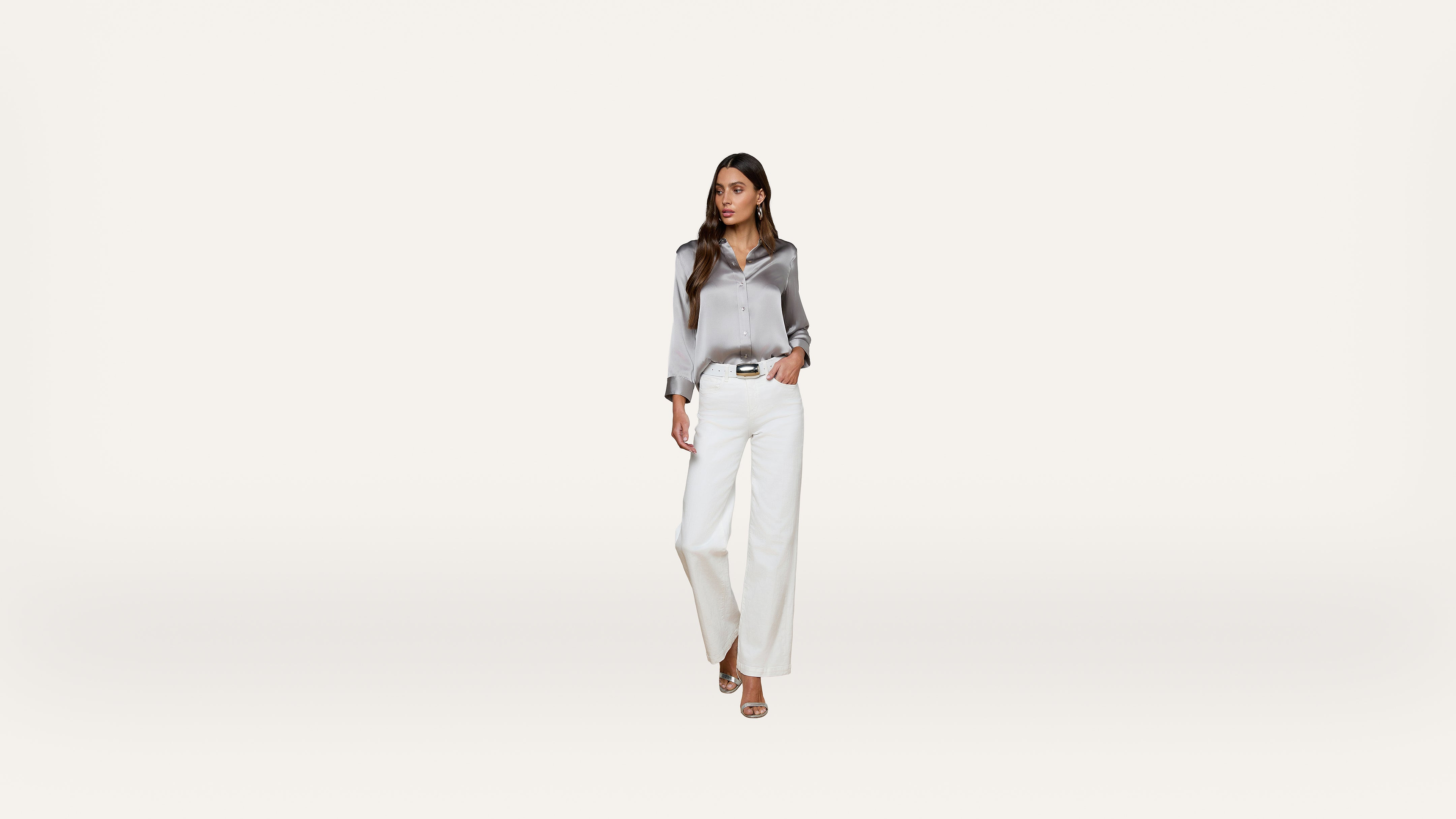 A woman wearing a shiny silver blouse, white wide-leg pants, and beige heels stands against a plain white background, looking to the side.