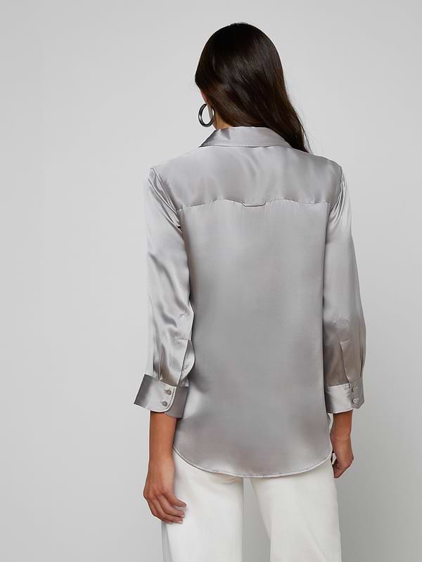 A woman with long dark hair, wearing the Dani Silk Blouse—a shiny silver silk button-down with long sleeves—paired with white pants, stands facing away from the camera against a plain light background.