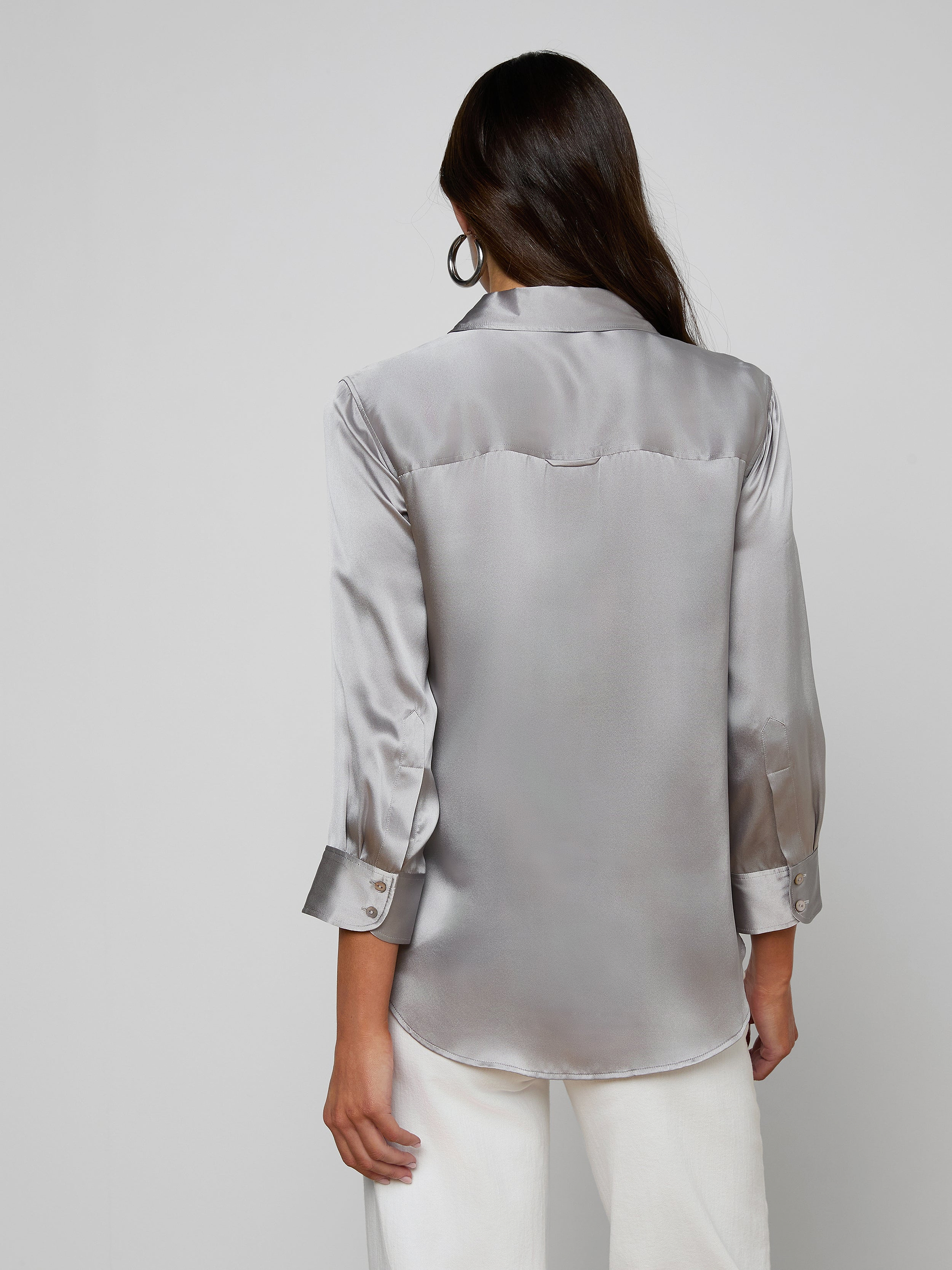 A woman with long dark hair, wearing the Dani Silk Blouse—a shiny silver silk button-down with long sleeves—paired with white pants, stands facing away from the camera against a plain light background.