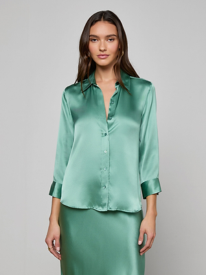 A woman with long brown hair wears the Dani Silk Blouse, a shiny light green 100% silk charmeuse top with buttons and 3/4 sleeves, standing against a plain light gray background.