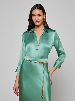 A woman poses against a plain light background, looking at the camera with her hand on her hip, wearing a shiny light green Dani Silk Blouse and skirt, styled with a gold chain belt.