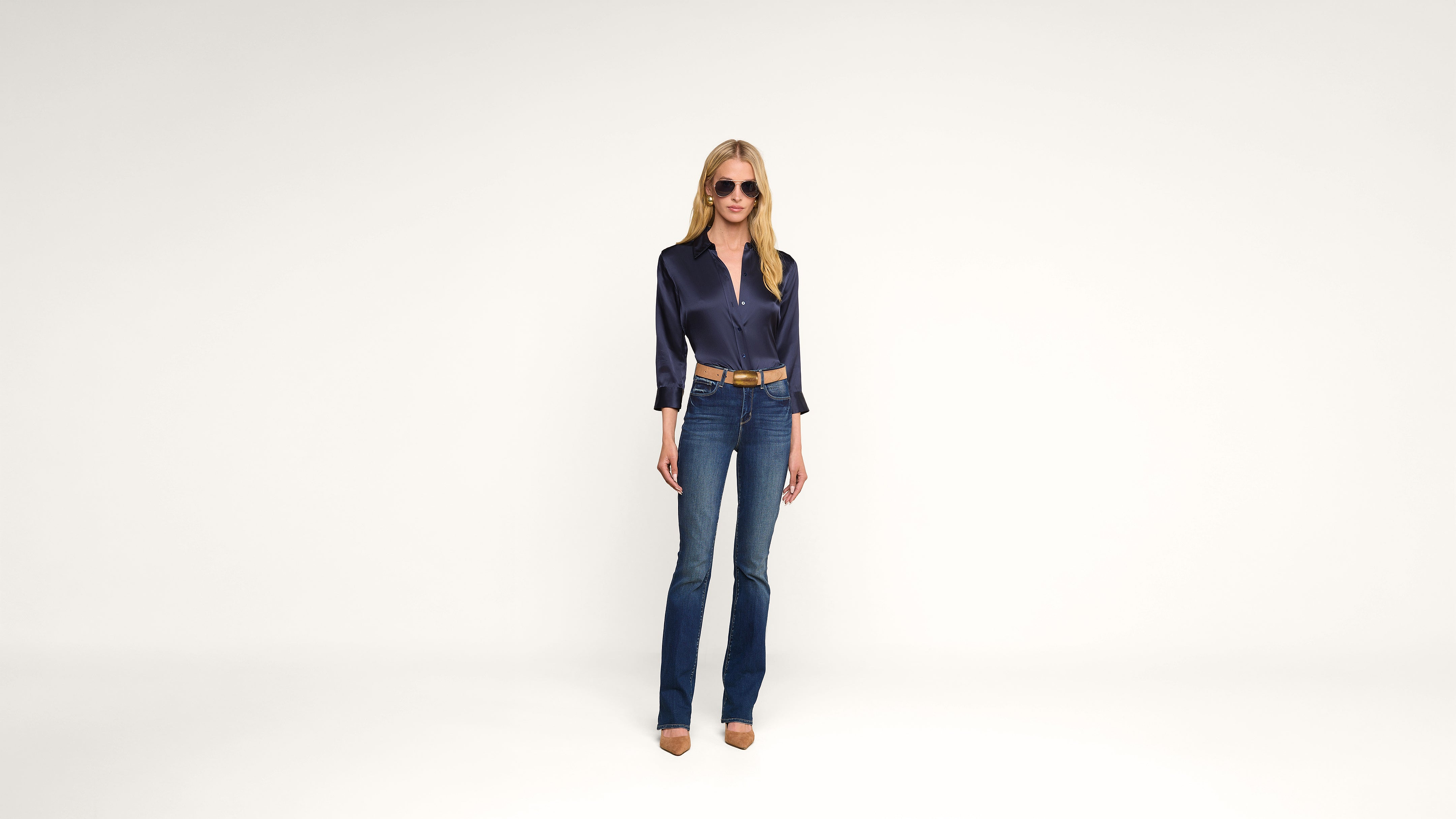 Woman stands against a plain white background wearing sunglasses, a dark blue satin blouse, blue flared jeans, a brown belt, and brown pointed shoes. She has long blonde hair and a relaxed posture.