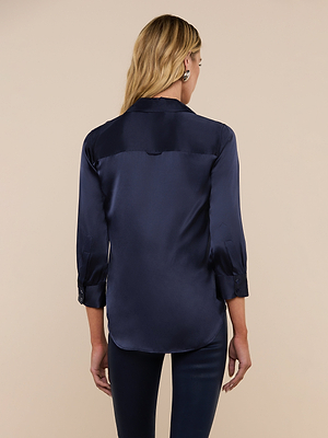 A woman with long blonde hair is seen from behind wearing the Dani Silk Blouse in midnight blue with matching dark pants, standing against a beige background.