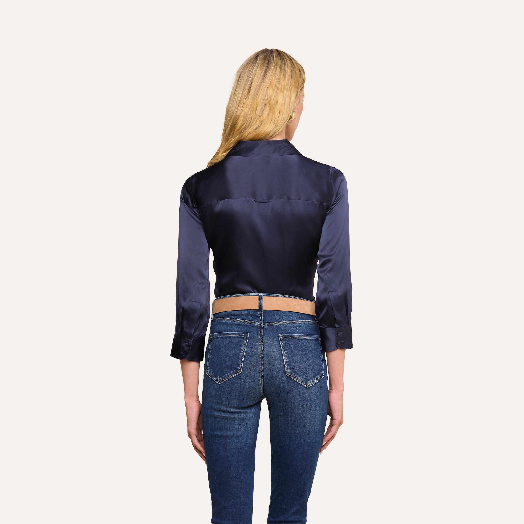 Eine Frau mit langen blonden Haaren ist von hinten zu sehen. Sie trägt die Dani-Seidenbluse in Mitternachtsblau, deren Ärmel hochgekrempelt sind, dazu eine blaue Jeans. Sie steht vor einem schlichten beigen Hintergrund.