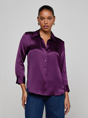 A woman wears the L'AGENCE Dani Silk Blouse in shiny deep purple with three-quarter sleeves and blue jeans, standing against a plain light gray background.