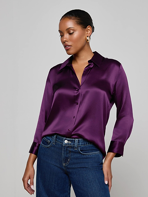 A woman wears the L'AGENCE Dani Silk Blouse in shiny purple with three-quarter sleeves, paired with dark blue jeans. She poses against a light gray background, accessorized with gold hoop earrings and her hair pulled back.