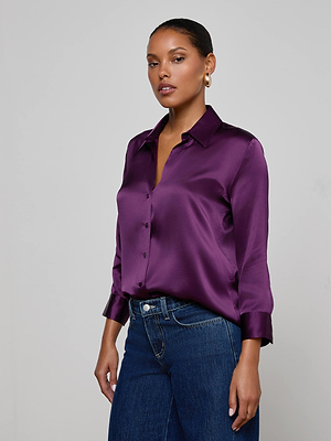 A woman models the L'AGENCE Dani Silk Blouse in shiny purple charmeuse with long sleeves, paired with dark blue jeans. She stands against a light gray background, hair pulled back, accessorized with gold hoop earrings.