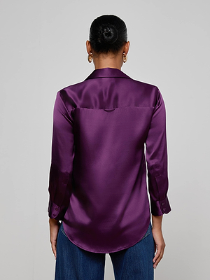 A person with their hair in a bun is seen from the back, wearing the L'AGENCE Dani Silk Blouse in shiny purple, paired with blue jeans against a plain light background.