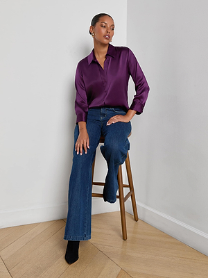 A person sits on a wooden stool by a white wall, wearing the L'AGENCE Dani Silk Blouse in purple with blue flared jeans and black shoes, one hand on their knee and the other resting on the stool.
