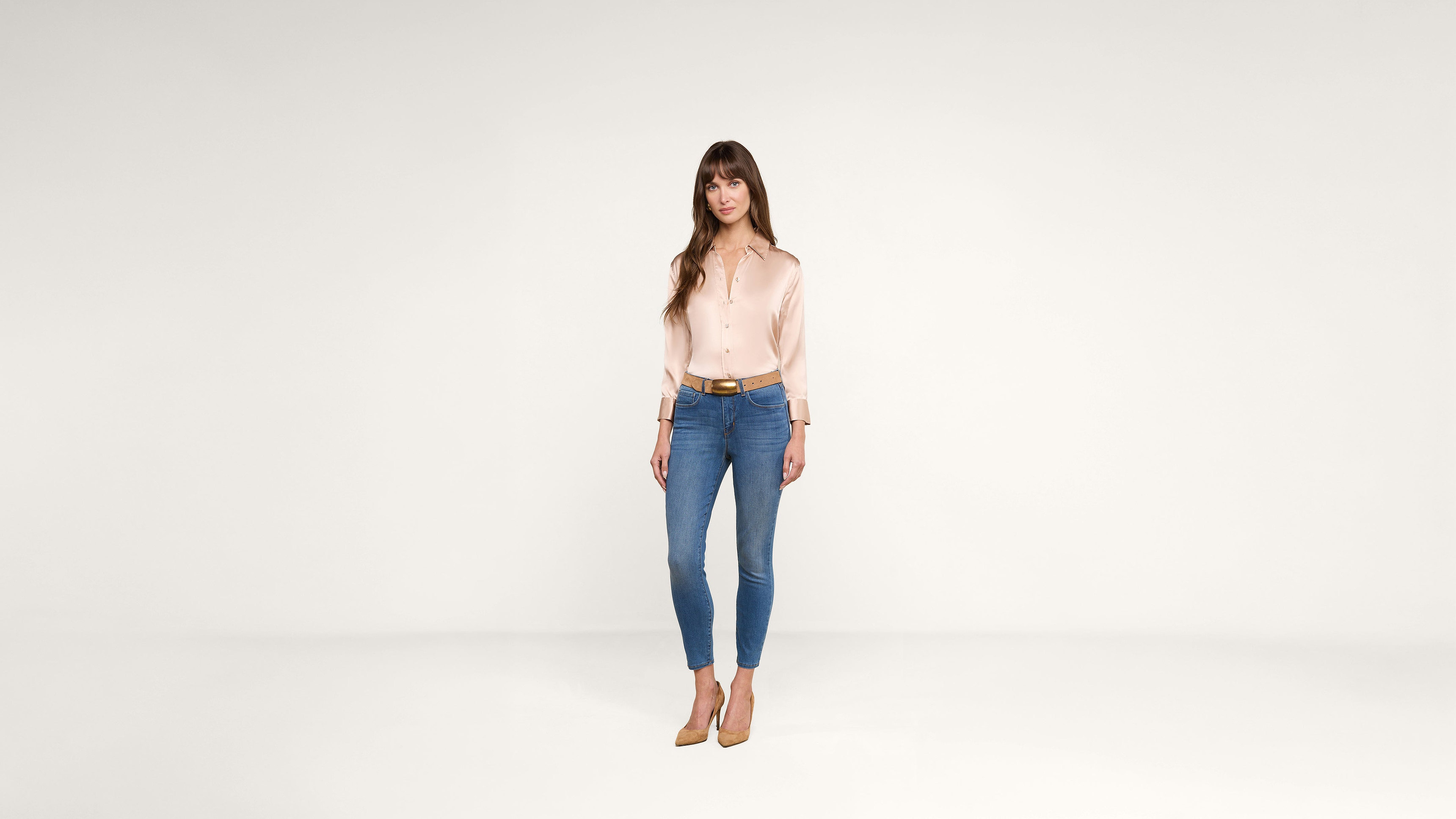 A woman with long brown hair, wearing a light pink blouse, blue jeans, and tan heels, stands facing forward against a plain white background.