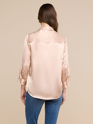Una mujer con el pelo largo y castaño lleva la blusa «Dani Silk», confeccionada en charmeuse de seda brillante de color beige claro, y unos vaqueros azules; aparece de espaldas a la cámara, con un fondo liso de color beige.