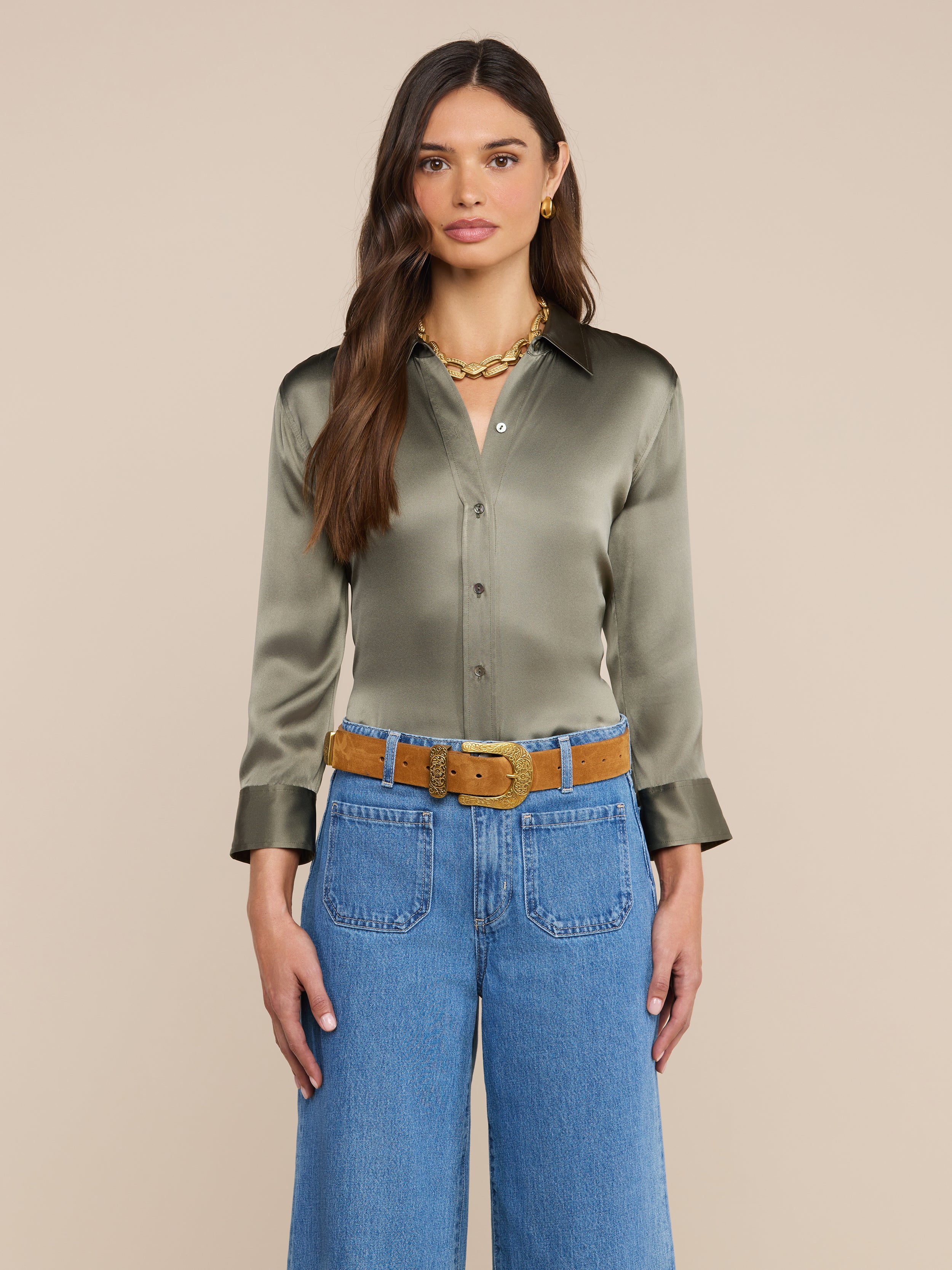 Vor einem beigen Hintergrund steht eine Frau mit langen braunen Haaren. Sie trägt eine raucholivgrüne Dani-Seidenbluse, eine blaue Jeans mit weitem Bein, einen hellbraunen Gürtel, goldene Creolen und eine klobige goldene Halskette.