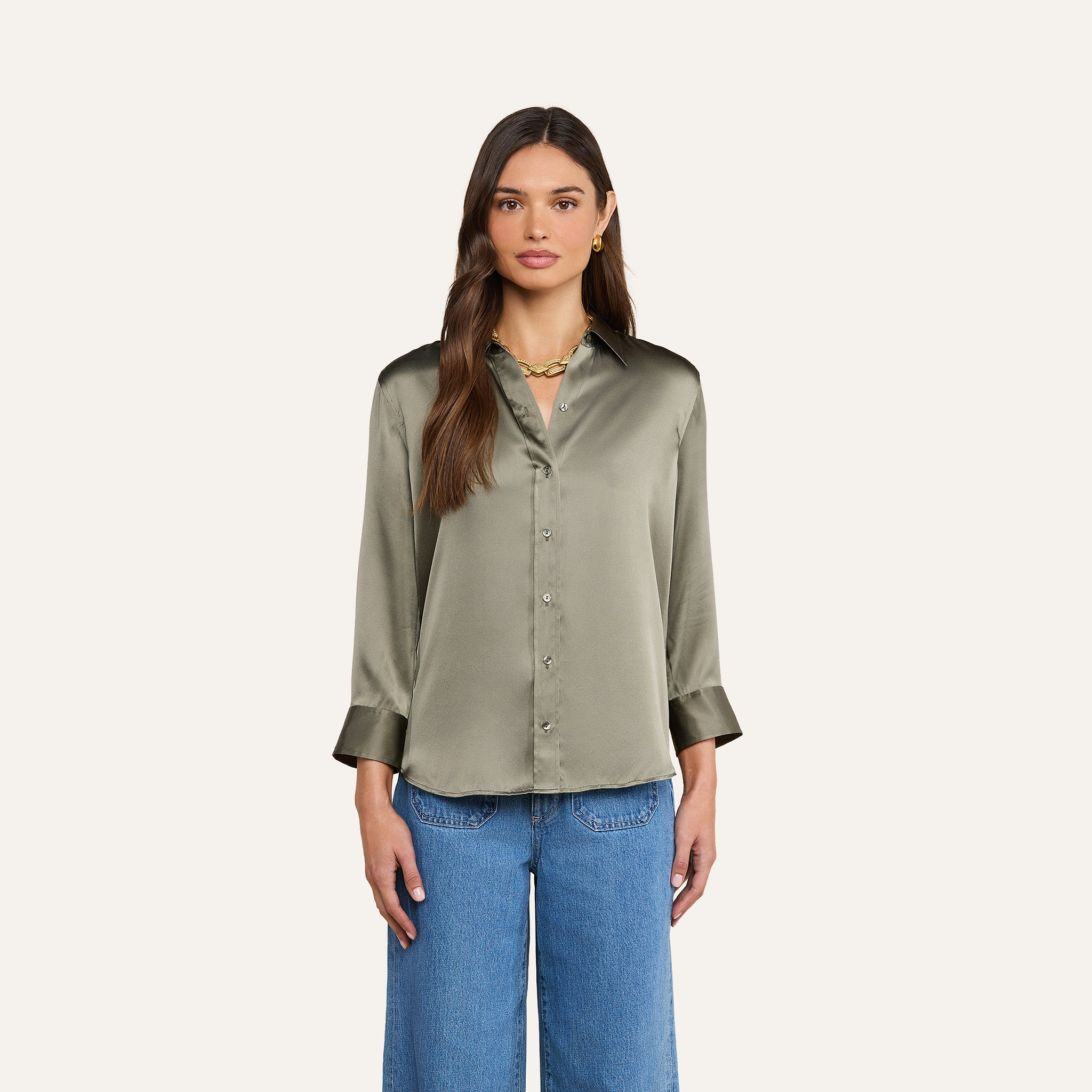 Eine Frau mit langen braunen Haaren trägt eine olivgrüne Dani-Seidenbluse in Smoky-Optik und eine blaue Jeans und steht vor einem schlichten beigen Hintergrund.