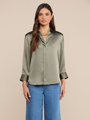 A woman with long brown hair wears a smoky olive green Dani Silk Blouse and blue jeans, standing against a plain beige background.
