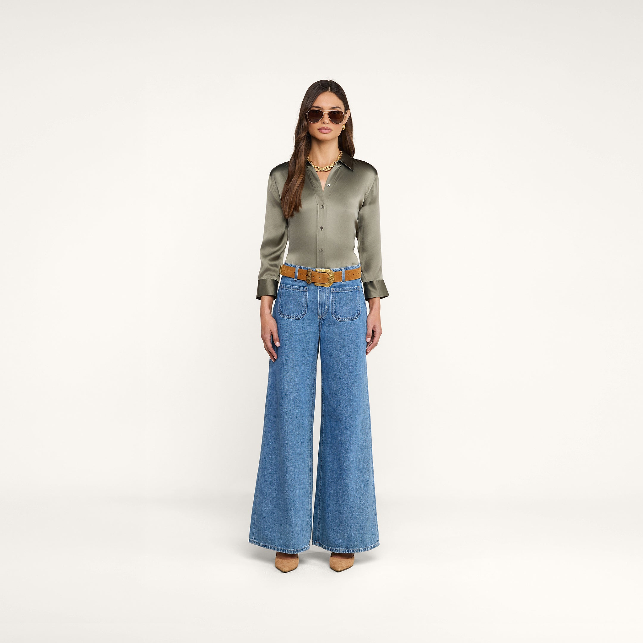 Vor einem schlichten beigen Hintergrund blickt eine Frau nach vorne. Sie trägt eine Sonnenbrille, eine rauchfarbene, olivgrüne Dani-Seidenbluse, eine blaue Jeans mit weitem Bein und einem braunen Gürtel, beige Schuhe mit Absatz sowie mehrere goldene Halsketten übereinander.