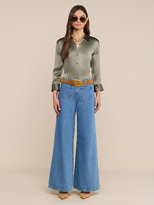 A woman faces forward, wearing sunglasses, a smoky olive green Dani Silk Blouse, wide-leg blue jeans with a brown belt, beige heeled shoes, and layered gold necklaces on a plain beige background.