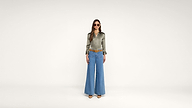 A woman stands against a plain white background, wearing sunglasses, a shiny green long-sleeve blouse, wide-leg blue jeans, a brown belt, and brown shoes.