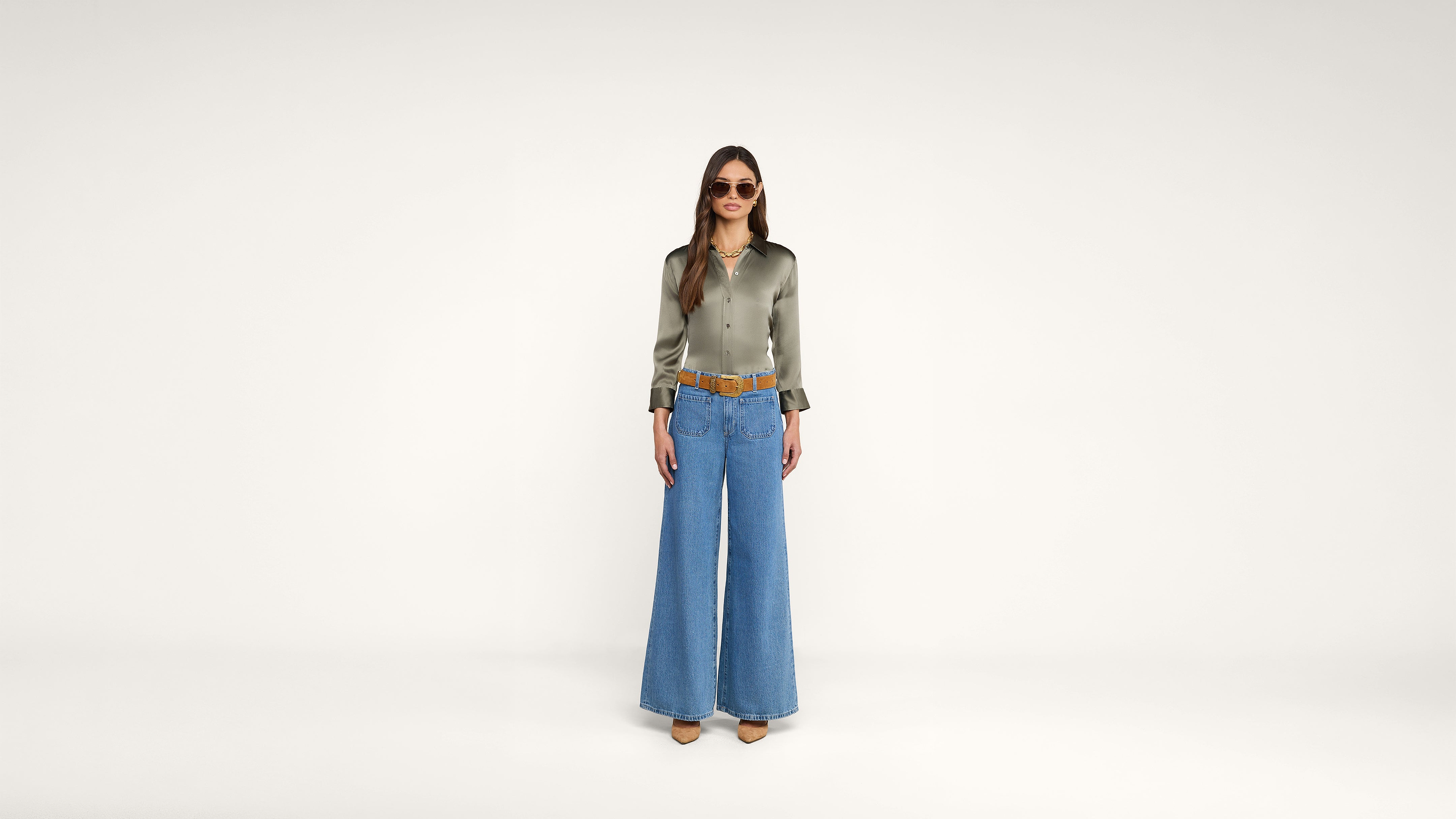 A woman stands against a plain white background, wearing sunglasses, a shiny green long-sleeve blouse, wide-leg blue jeans, a brown belt, and brown shoes.