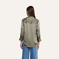 Vor einem beigen Hintergrund ist eine Frau mit langen braunen Haaren von hinten zu sehen, die die Dani-Seidenbluse in rauchigem Olivgrün und eine blaue Jeans mit weitem Bein trägt.