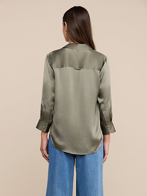 A woman with long brown hair is seen from behind wearing the Dani Silk Blouse in smoky olive green and blue wide-leg jeans against a beige background.