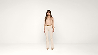 A woman stands in the center of a plain white background, wearing a beige blouse, cream pants, brown belt, and brown shoes. She faces forward with her arms relaxed by her sides.