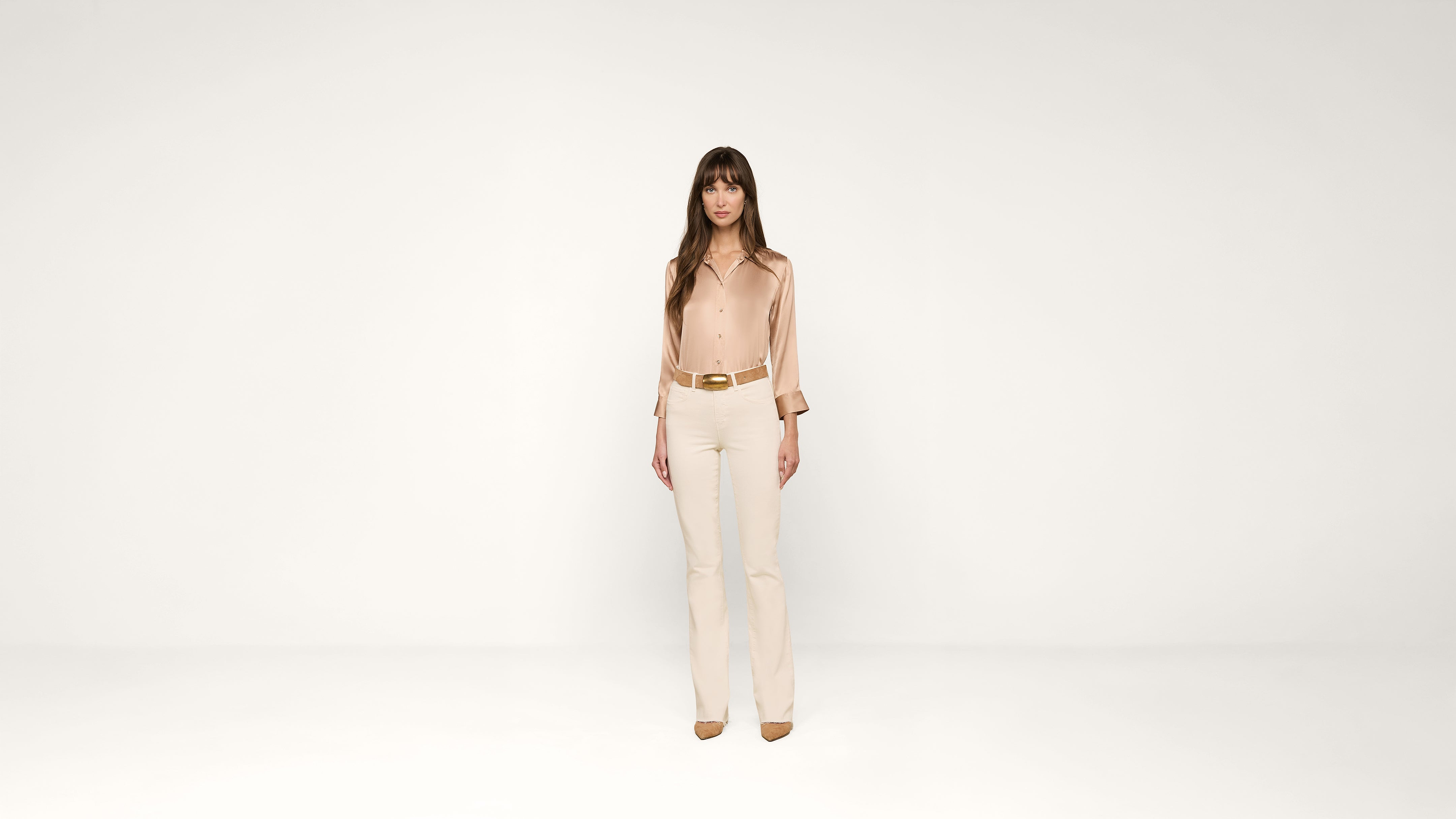A woman stands in the center of a plain white background, wearing a beige blouse, cream pants, brown belt, and brown shoes. She faces forward with her arms relaxed by her sides.