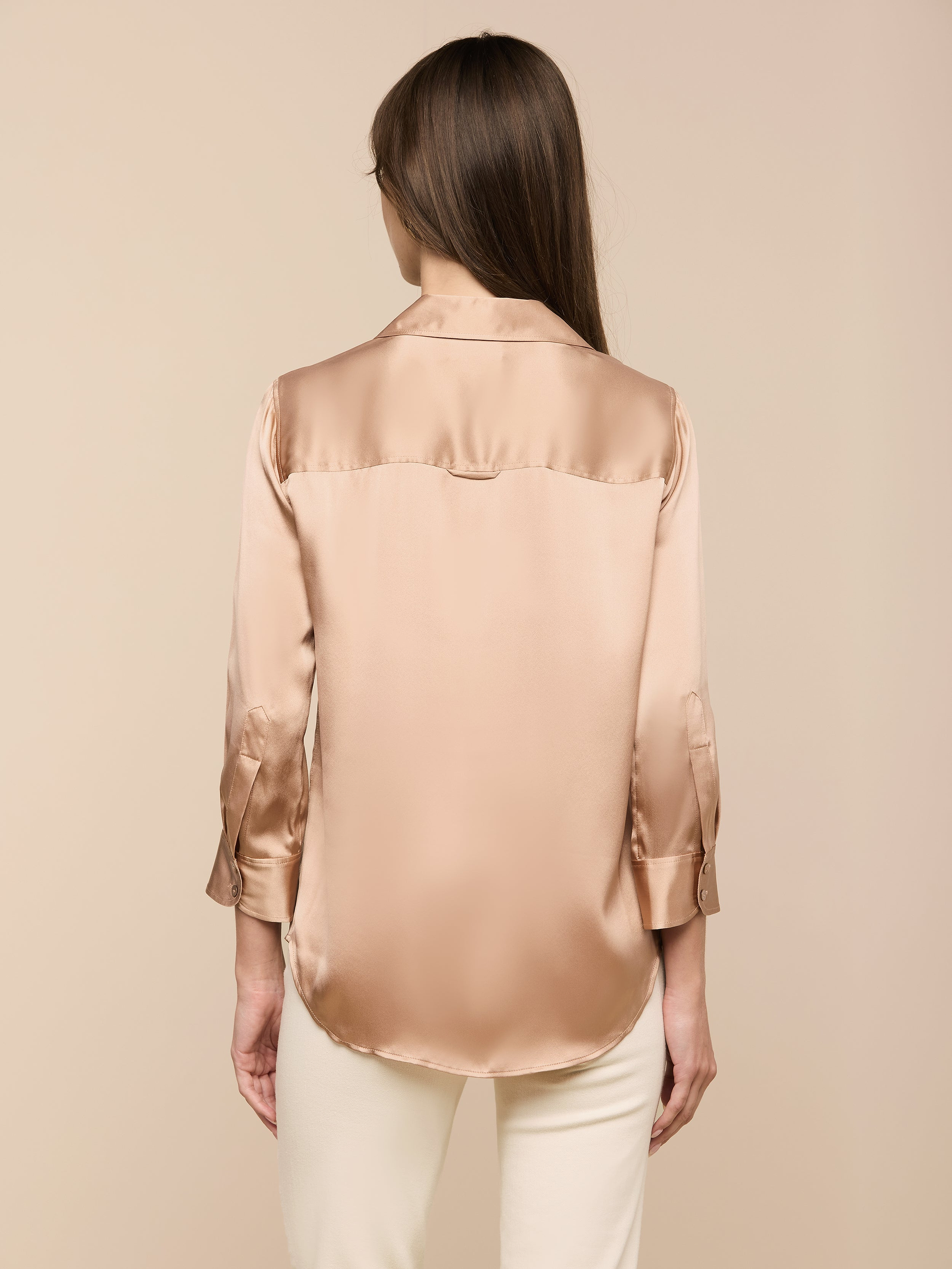 A woman with straight brown hair, seen from behind, wears the Dani Silk Blouse—a shiny, light brown, long-sleeve silk top—paired with white pants and standing against a plain, light background.