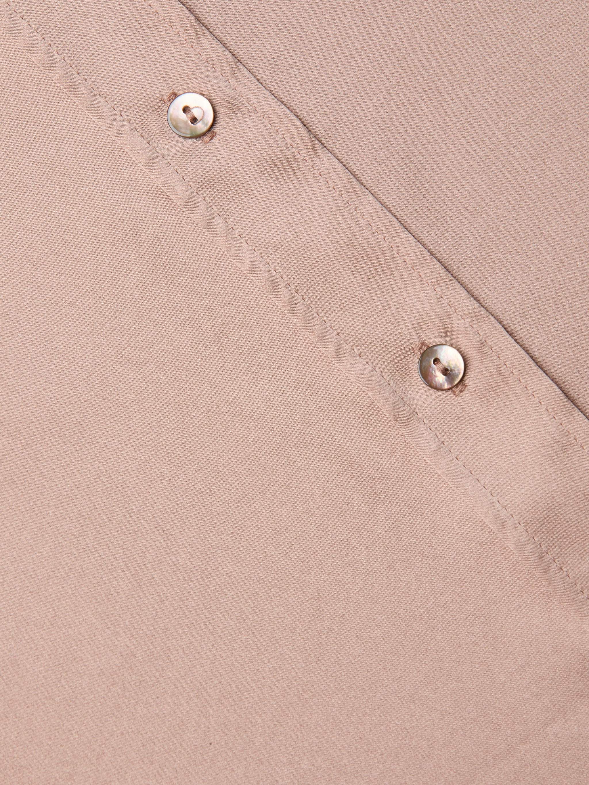 Close-up of the Dani Silk Blouse in light brown, featuring a diagonal stitched seam and two round, shiny buttons along the seam. The smooth texture of the silk material is visible.