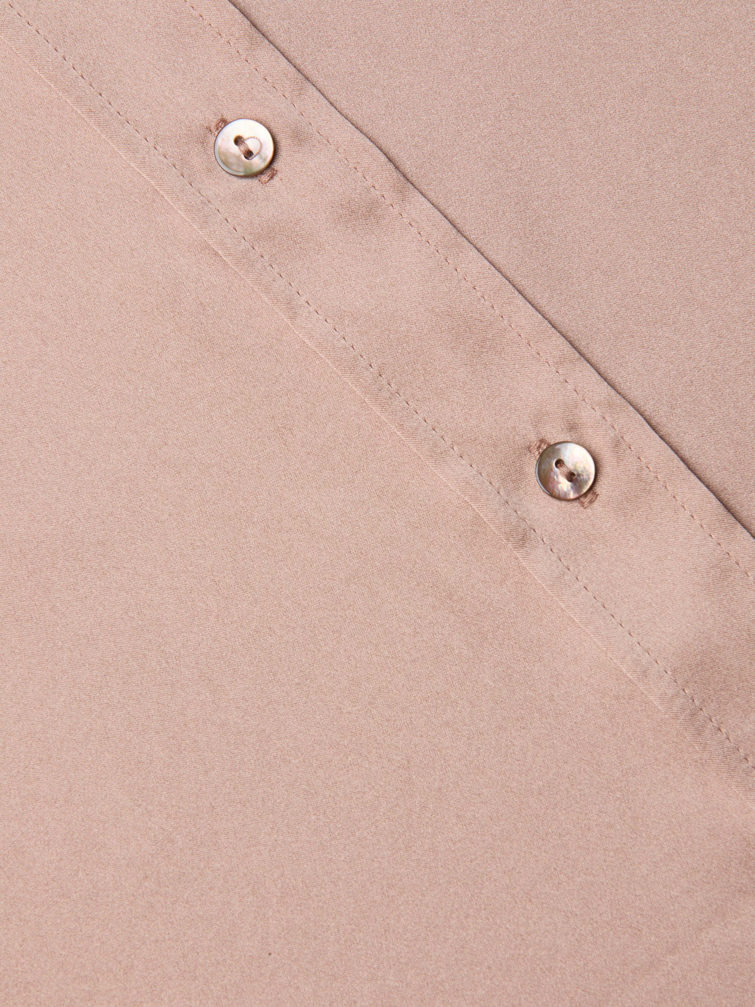 Close-up of the Dani Silk Blouse in light brown, featuring a diagonal stitched seam and two round, shiny buttons along the seam. The smooth texture of the silk material is visible.
