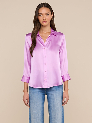 A woman with long brown hair wears the Dani Silk Blouse, a shiny light purple button-down with three-quarter sleeves, paired with light blue jeans, standing facing forward against a plain beige background.
