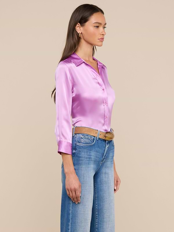 A woman stands in profile wearing the shiny pink Dani Silk Blouse, styled with blue jeans and a brown belt. She has long brown hair and looks ahead against a plain light background.