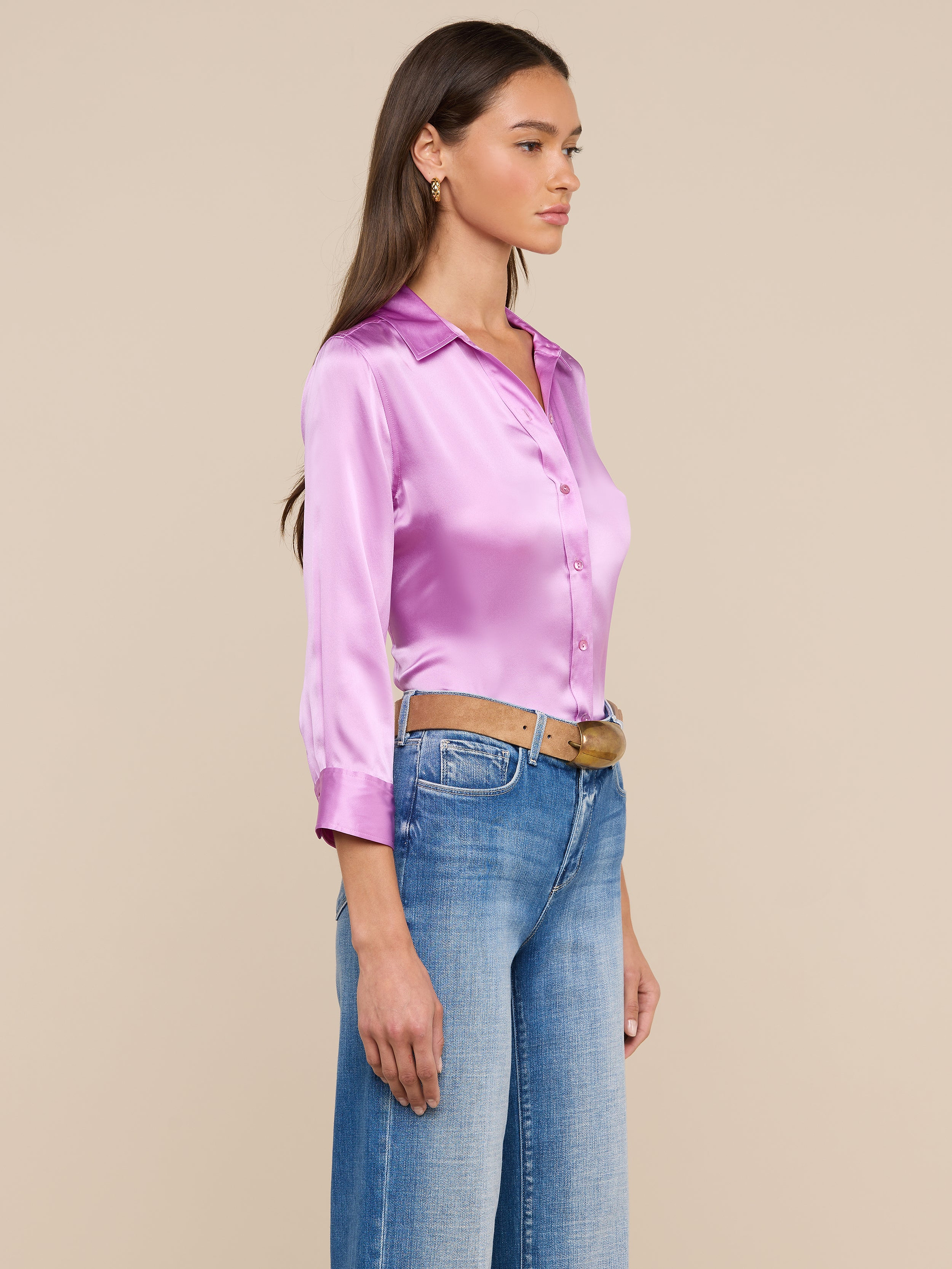 A woman stands in profile wearing the shiny pink Dani Silk Blouse, styled with blue jeans and a brown belt. She has long brown hair and looks ahead against a plain light background.