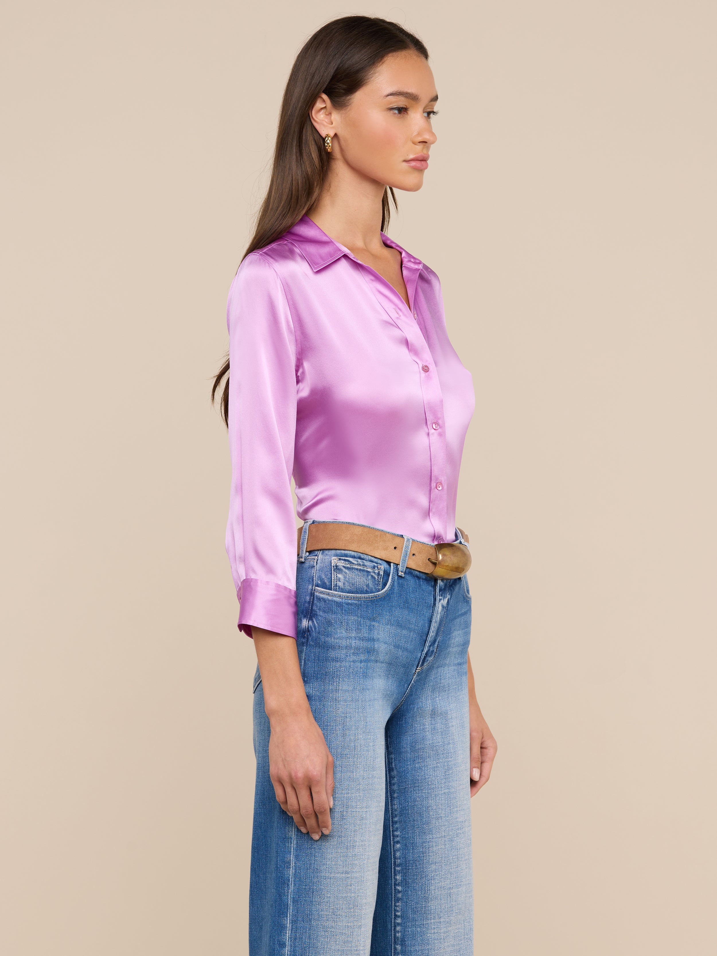 A woman stands in profile wearing the shiny pink Dani Silk Blouse, styled with blue jeans and a brown belt. She has long brown hair and looks ahead against a plain light background.