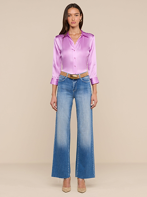 A woman with long hair and a neutral expression stands against a beige background wearing the Dani Silk Blouse in shiny lavender, three-quarter sleeves, wide-leg blue jeans with a brown belt, and beige heels.