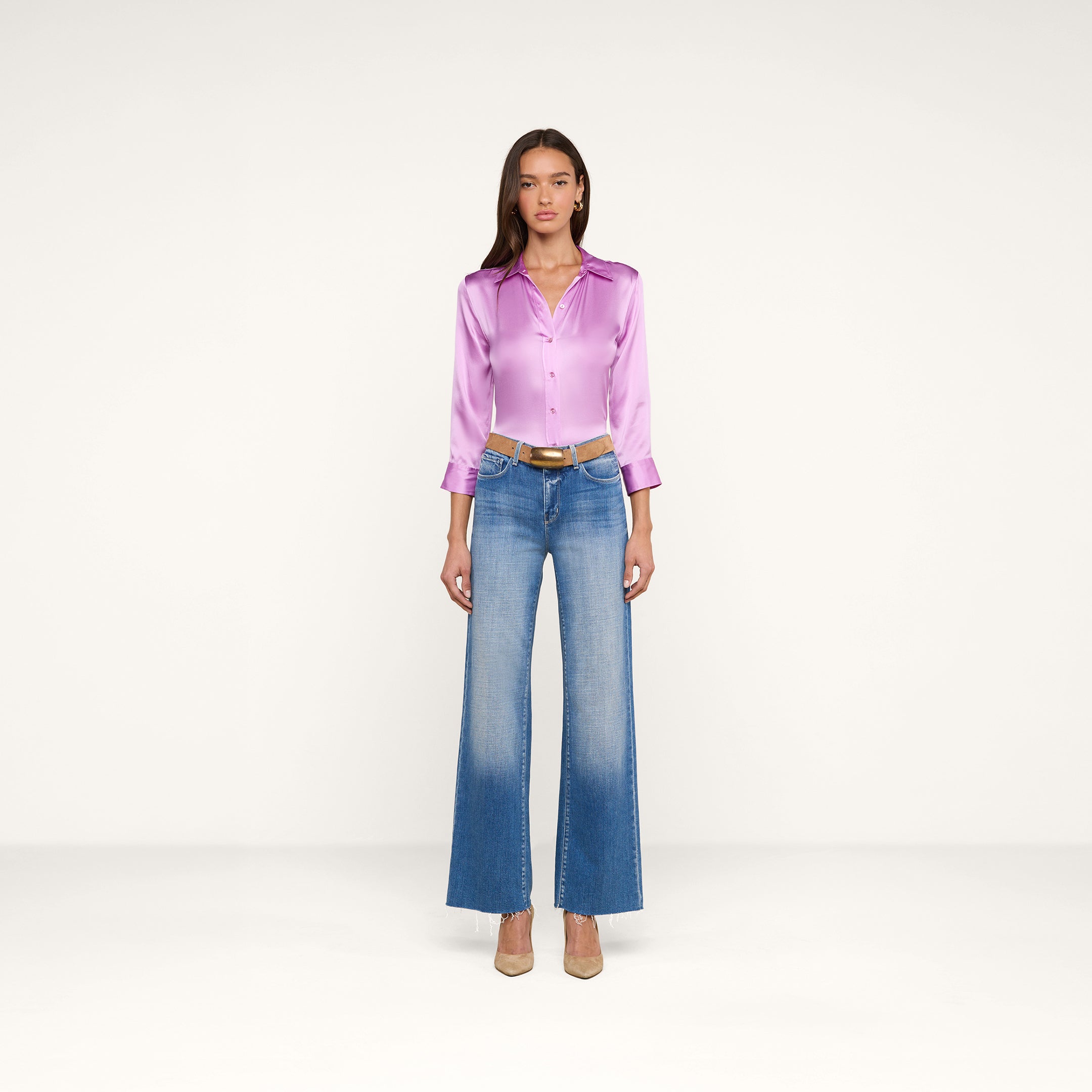 A woman stands facing forward against a plain white background, wearing the shiny lavender Dani Silk Blouse in silk charmeuse, blue high-waisted wide-leg jeans, a brown belt, and beige heels.