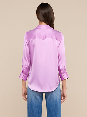 A person with long brown hair is shown from behind, wearing the Dani Silk Blouse in shiny light purple with three-quarter sleeves and light blue jeans against a plain beige background.